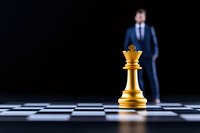 Golden queen chess piece on board with blurred businessman in background, representing leadership strategy and business competition concept.