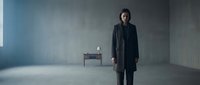 Young adult male with dark hair and East Asian ethnicity wearing a black formal coat standing in an empty industrial concrete space with minimal furniture, suitable for corporate, fashion, and editorial projects.