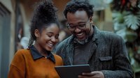 Young African American couple smiling while looking at digital tablet outdoors. Woman wearing orange sweater and man in glasses and jacket sharing joyful moment.