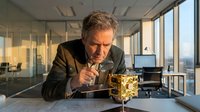 Mature businessman with gray hair and facial hair, Caucasian ethnicity, examining golden coins at desk in modern high-rise office with city skyline view, representing wealth analysis and financial success.