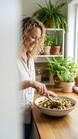 Happy middle-aged Caucasian woman preparing healthy quinoa salad in modern kitchen with houseplants on shelves for nutritious home cooking lifestyle.