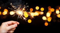 Hand holding burning sparkler with golden sparks against blurred bokeh lights background for celebration and festive occasions.