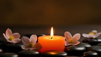 Orange candle burning with soft flame surrounded by pink cherry blossoms and smooth black spa stones creating peaceful zen meditation atmosphere.