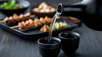 Pouring sake into traditional black ceramic cups with grilled yakitori skewers and side dishes on dark wooden table, Japanese dining experience.