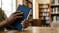 Senior man with dark skin tone holding closed navy blue book at wooden table in library with bookshelves, reading and education concept for publications and learning materials.