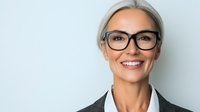 Mature businesswoman with silver hair wearing glasses and professional black blazer smiling confidently against light background. Professional corporate headshot.