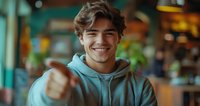 Young Caucasian man in mint hoodie showing thumbs up with confident smile against blurred cafe background, expressing positivity and approval.