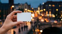 Close-up of hand holding blank white card against evening cityscape with bokeh lights, suitable for business cards, travel promotions, and urban marketing campaigns.