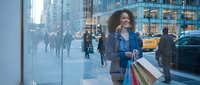 Young African American woman with curly hair enjoying shopping in busy urban city street, carrying colorful bags while talking on phone during evening.