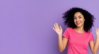 Young woman with curly black hair and medium skin tone wearing bright pink t-shirt, waving hands with cheerful expression against purple background for lifestyle and portrait use.