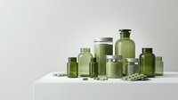 Green glass pharmaceutical bottles and jars collection with pills scattered on white surface for medical packaging and healthcare branding designs.