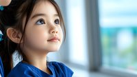 Young Asian girl in blue dress gazing upward with hopeful expression near window, natural daylight illuminating her face, capturing childhood wonder and dreams.