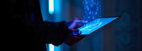 Glowing digital tablet with holographic data stream in dark room creating futuristic blue light effect, person holding device with emerging technology visualization.