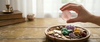 Hand holding clear quartz crystal above wooden bowl filled with colorful healing gemstones on rustic table for meditation and spiritual wellness practices.