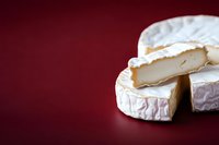 Soft ripened white mold cheese wheel cut into wedges on dark red background, showing creamy texture and characteristic rind. Professional food photography.
