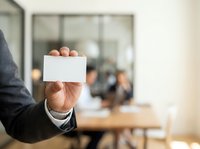 Close-up of male professional in dark suit holding blank white business card in office meeting room with blurred colleagues in background.