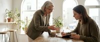 Senior businesswoman mentor coaching young female employee at modern office desk with plants and natural lighting for professional development.