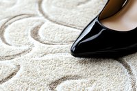 Elegant black patent leather high heel shoe on luxurious cream carpet with floral pattern, creating contrast between formal footwear and soft home decor texture.