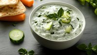 Fresh tzatziki sauce with diced cucumber, herbs and yogurt in white ceramic bowl on dark stone background, garnished with basil leaves and vegetables.