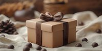 Kraft paper gift box with brown satin ribbon bow surrounded by chocolate candies and pine cones on soft beige textile background. Festive holiday concept.