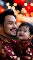 Asian father and young son wearing traditional red and gold attire share joyful moment against festive bokeh lights background during lunar new year celebration.