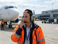 Female airport ground crew worker with headset communicating at busy commercial airport terminal with aircraft and ground support equipment.