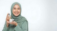 Young Muslim woman in green hijab holding skincare serum bottle, smiling at camera on white background for beauty and cosmetic advertising campaigns.