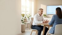 Middle-aged Caucasian man in business casual attire having a professional conversation with a young woman in modern office setting with natural light and plants.
