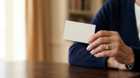 Mature man with medium skin tone wearing navy blazer holding blank white business card at wooden table, suitable for corporate identity, professional networking, and business communication concepts.