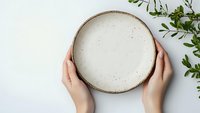 Female hands holding empty ceramic plate on white background with green plant decoration. Minimalist table setting concept for food photography and styling.