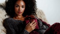 Young African American woman in cozy gray sweater holding burgundy gift box with bow, lying on fluffy blanket. Natural curly hair and soft winter holiday mood.