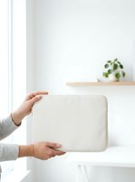 Woman holding cream laptop sleeve in bright minimalist interior with potted plant on shelf, suitable for tech product marketing and lifestyle content.