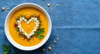 Creamy pumpkin soup with heart-shaped cream garnish and seeds on rustic blue wooden background. Healthy comfort food for autumn dining and seasonal cooking.