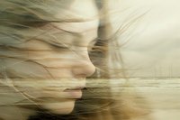 Double exposure portrait of young woman with blonde hair merged with serene coastal landscape, wind-blown hair creating dreamy atmospheric mood.