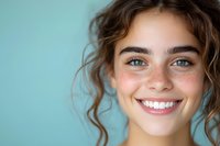 Close up portrait of young woman with natural freckles, curly brown hair and bright blue eyes smiling genuinely against turquoise background.