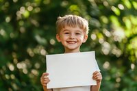 Cheerful young boy holding blank white paper outdoors in garden with natural bokeh background. Genuine smile and positive emotion for advertising or message.