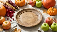 Wooden plate surrounded by autumn harvest produce including apples, pumpkins, corn and berries on white wooden surface for fall seasonal cooking and recipe content.