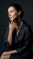 Elegant middle-aged Caucasian woman in silk robe applying dark mud mask to neck, spa skincare concept on dark background.