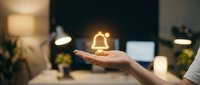 Hand holding glowing bell icon above desk with laptop and plants, representing notification, alert, customer service, and digital communication in modern office environment.