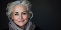 Elegant mature woman with silver gray hair and natural makeup smiling confidently against dark background, wearing cozy scarf, representing aging gracefully and vitality.
