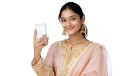 Young South Asian woman in traditional peach-colored salwar kameez with gold embroidery taking selfie on smartphone, wearing traditional jewelry and smiling at camera on white background.
