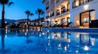 Luxurious resort swimming pool illuminated at dusk with reflective blue water, palm trees, and elegant white building facade featuring balconies and ambient lighting.