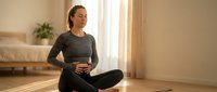 Young Asian woman practicing mindfulness meditation in lotus position on yoga mat in bright modern bedroom with natural sunlight streaming through window.