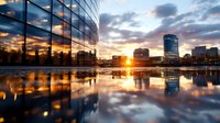 Modern glass office building with dramatic sunset reflection in urban puddle, cityscape with skyscrapers silhouettes against golden sky and clouds.
