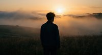 Young man silhouette standing in misty mountain landscape at golden sunrise, contemplating peaceful morning view with fog rolling over hills and valleys.