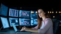 Young Caucasian woman working at night on laptop with multiple monitors displaying data analytics in dark office environment.