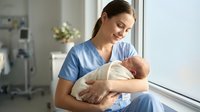 Young Caucasian female nurse in blue scrubs gently holding newborn baby in bright hospital room, maternal care and healthcare professional bonding moment.