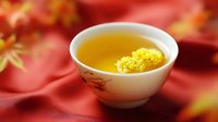 Yellow chrysanthemum flower floating in traditional Asian ceramic tea cup with hot tea against red silk background with autumn maple leaves.