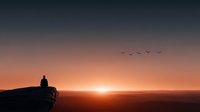 Silhouette of person sitting on rocky cliff edge watching sunrise over mountain landscape with flying birds in colorful sky for meditation and travel content.