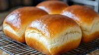 Freshly baked golden milk bread rolls cooling on wire rack, showing soft fluffy texture and glossy top surface. Close up view of homemade Japanese style bread.